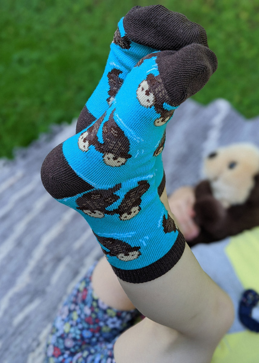 A child model wearing otter-themed novelty socks poses with their feet up in the air while holding a stuffed otter and laying in the grass.