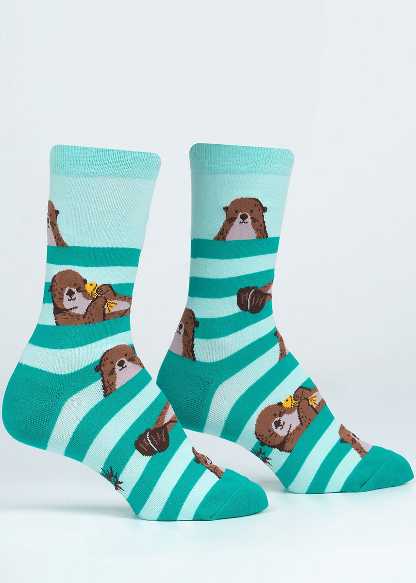 Cute animal socks for women show otters holding a yellow fish and hanging out among a background of teal stripes.