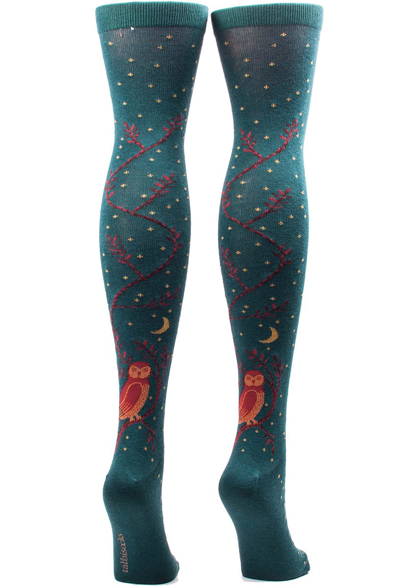Over-the-knee socks for women feature tiny gold stars, an owl, and the moon on a dark teal background on the back of your legs.