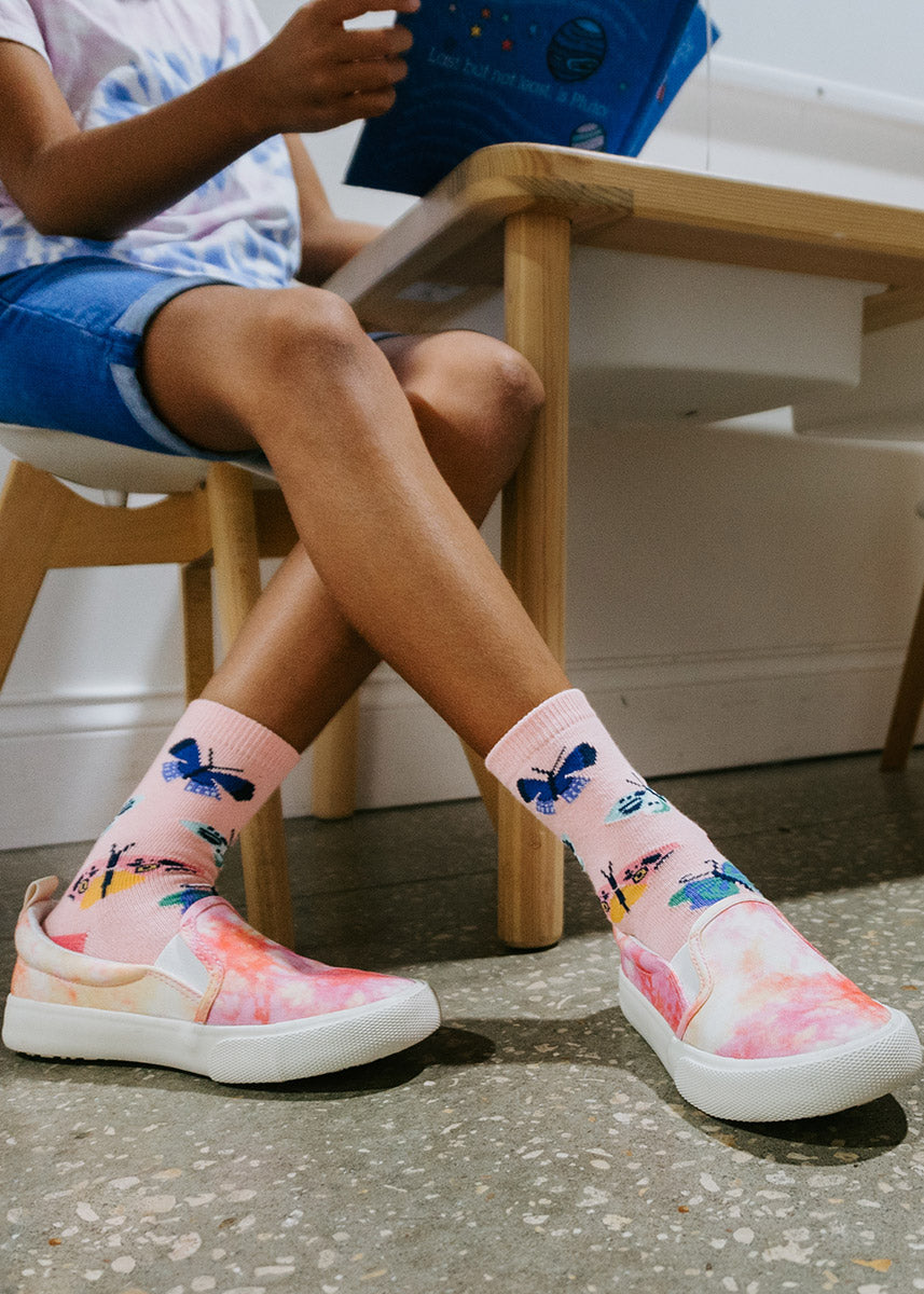 A female child model wearing butterfly-themed novelty socks poses sitting at a desk and reading a book.