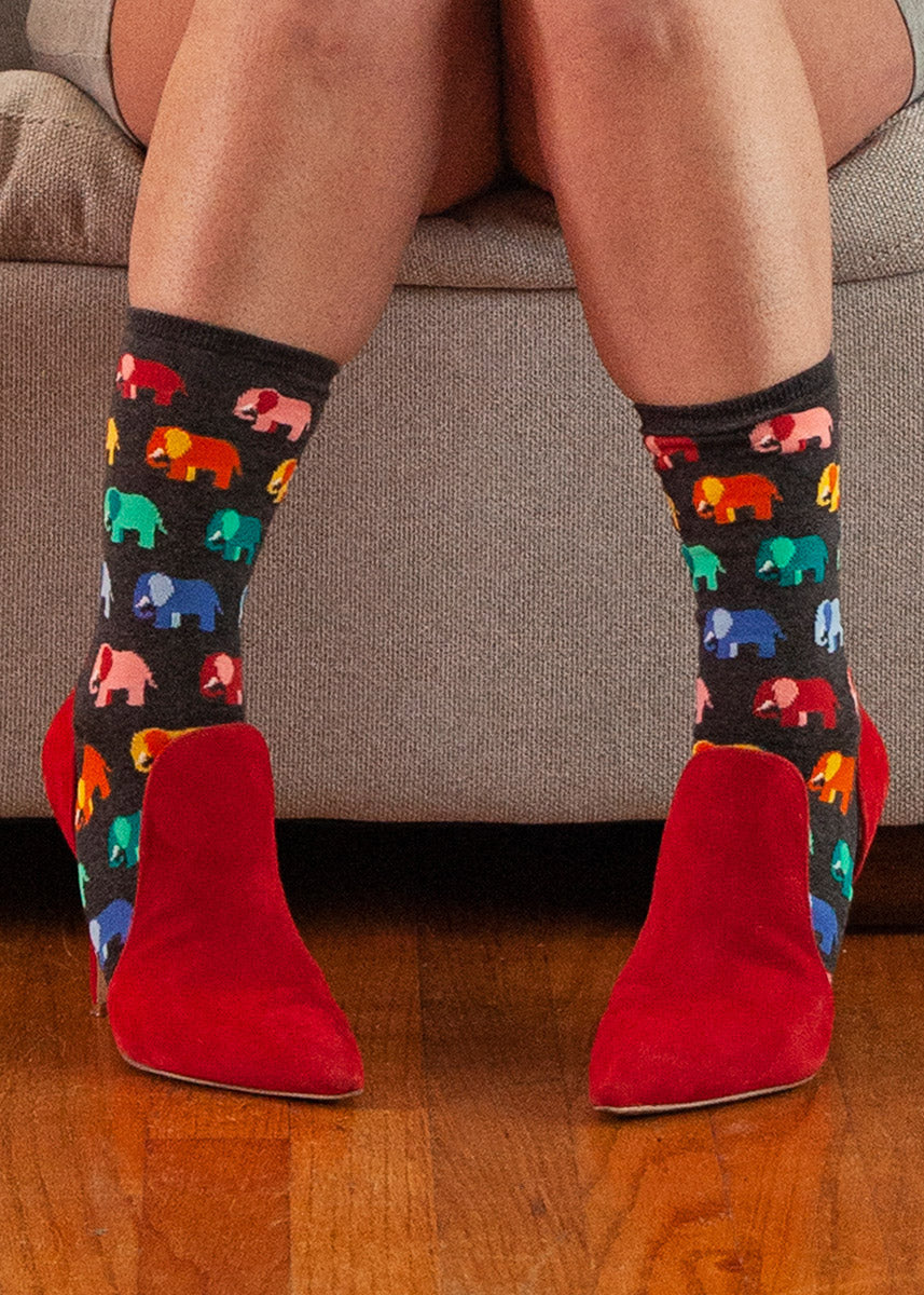 A model wearing rainbow elephant-themed novelty socks and red shoes poses seated on a couch
