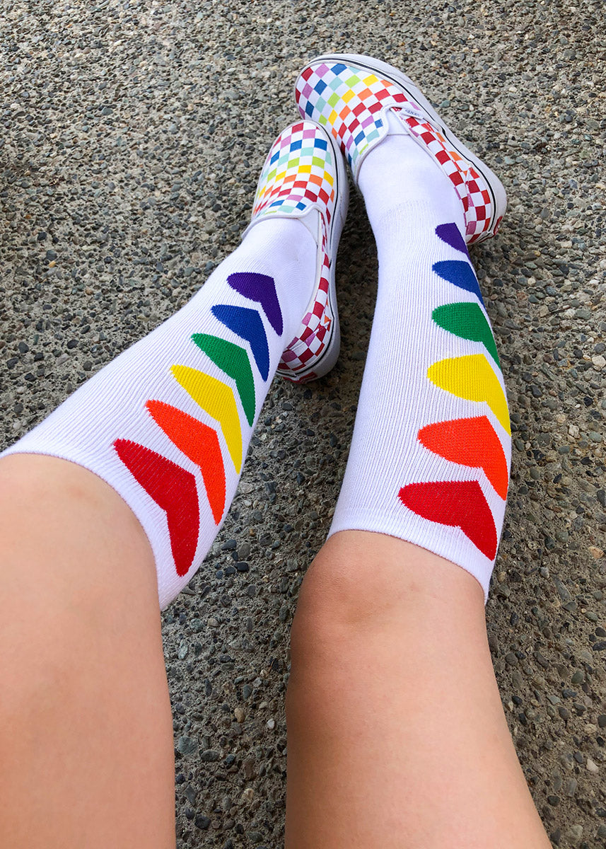 A model sits with legs outstretched, wearing rainbow checkered vans , and white knee high socks with individual rainbow hearts in a line on the sides of the sock.