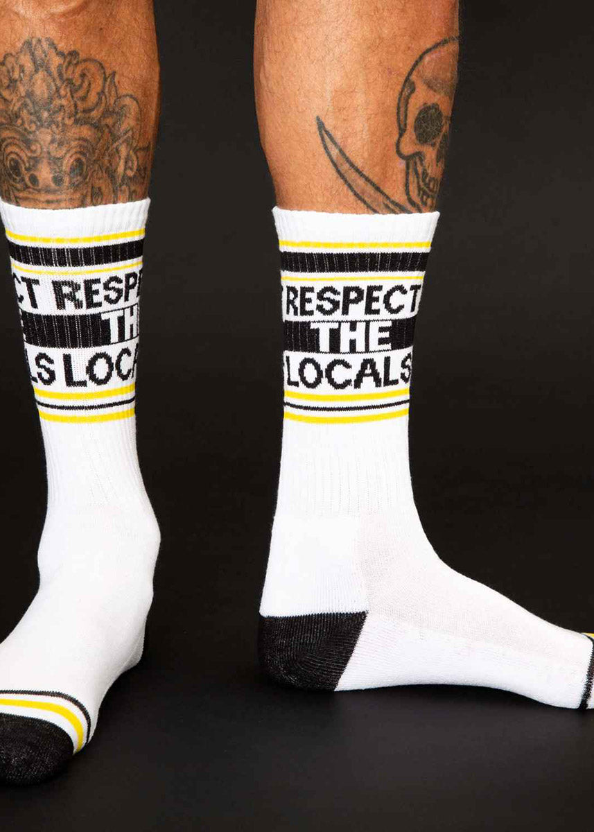 A tattooed male model poses shoeless against a black background wearing novelty socks that say &quot;RESPECT THE LOCALS.&quot;