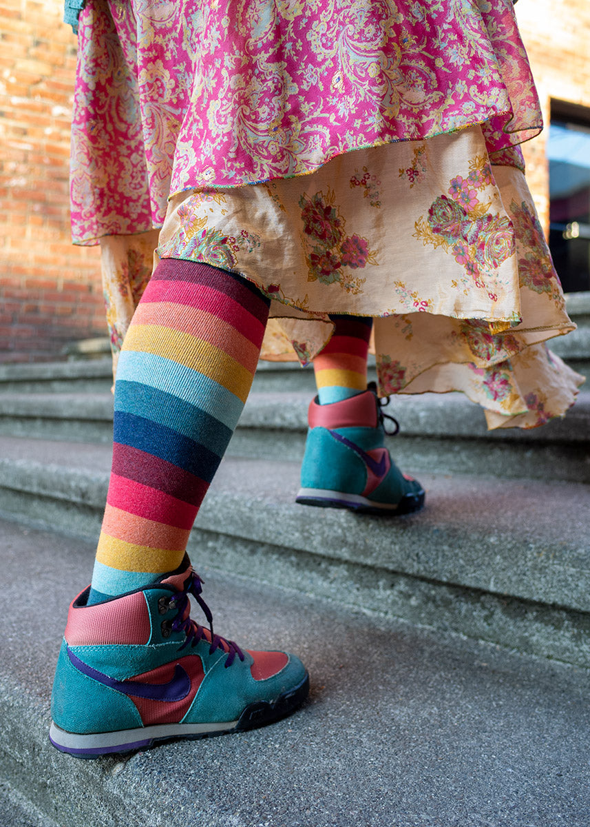 A female model wearing colorful striped knee socks and colorful sneakers poses walking up a set of concrete steps.