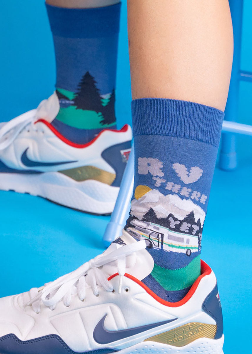 Blue men's crew socks with RVs driving down the open road with the words “RV There Yet?” written in clouds of exhaust, all with a background of trees and mountains.