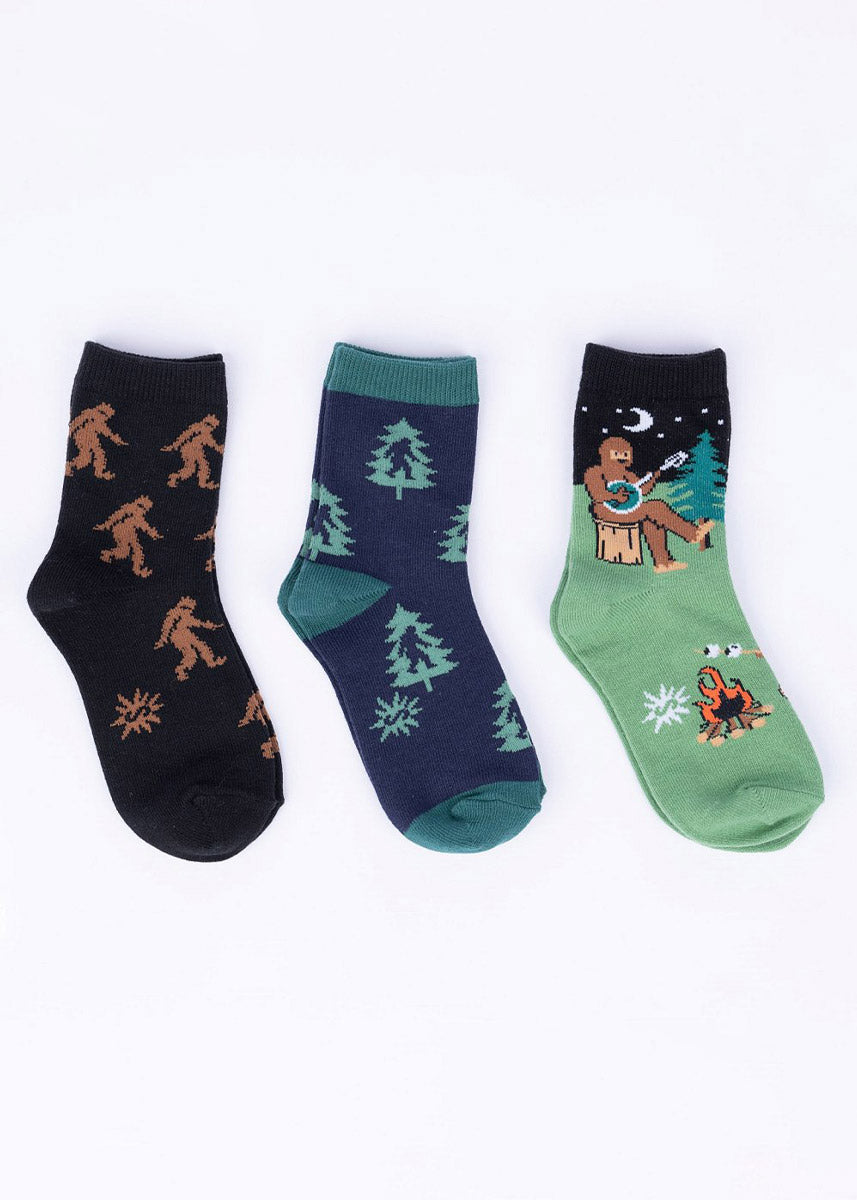 Three different pairs of coordinating Sasquatch-themed socks for kids including Bigfoot, evergreen forest and camping motifs.
