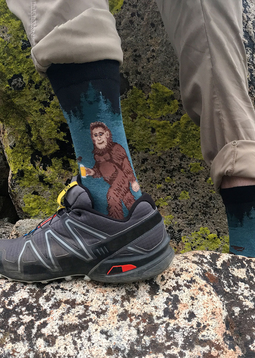 A male model wearing Sasquatch and beer-themed novelty socks and hiking shoes poses outside on a rock.