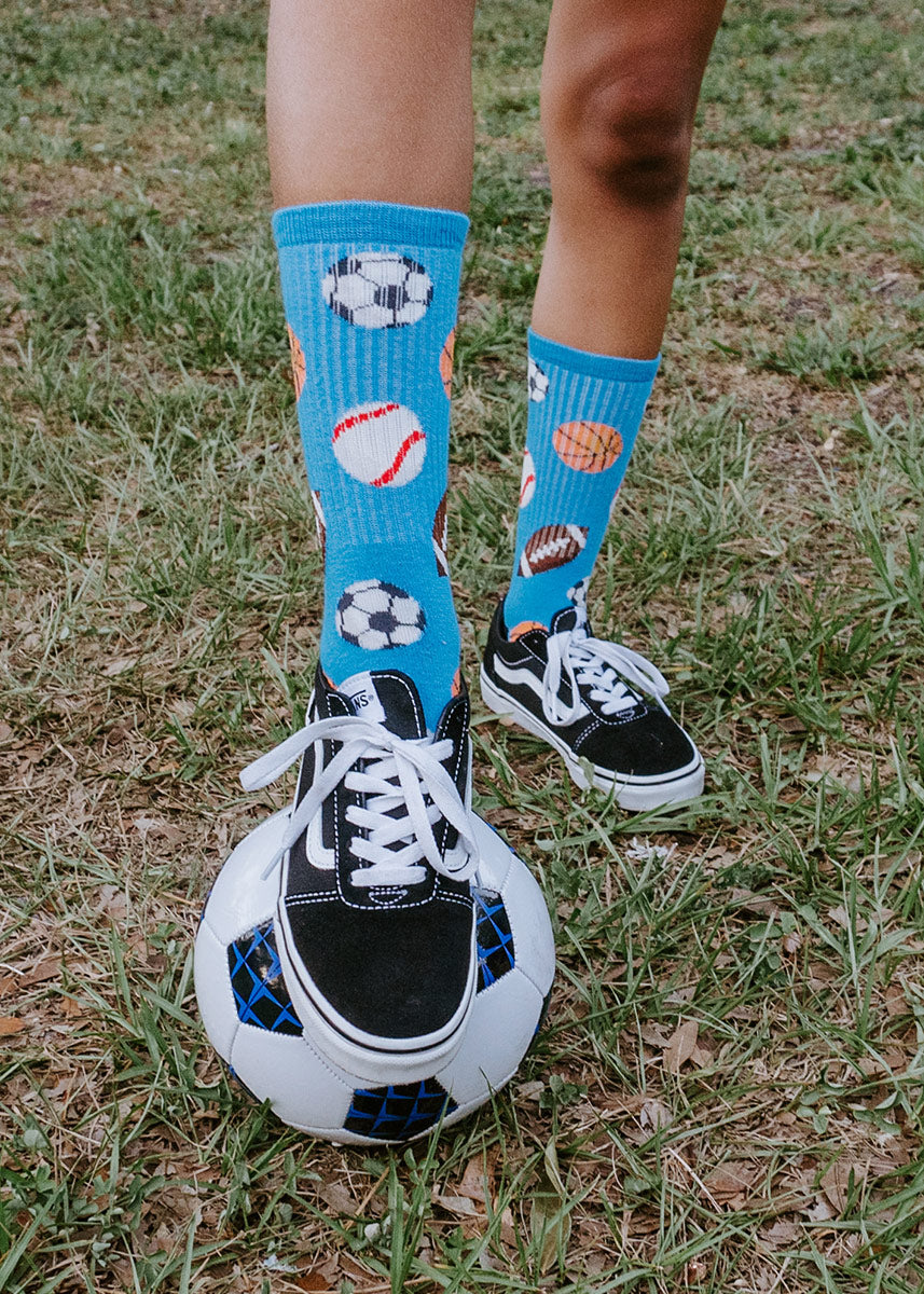 A child model wearing sports ball-themed novelty socks and black sneakers poses with a soccer ball in a grass field. 