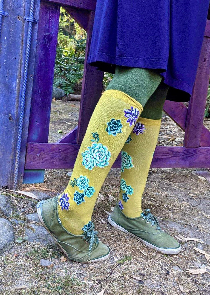 A model wearing green succulent-themed novelty knee socks poses outside next to a wooden gate. 