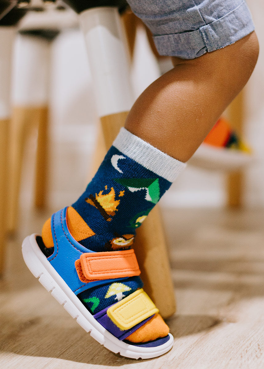 A child model wearing camping-themed novelty socks and colorful sandals poses seated on a chair.