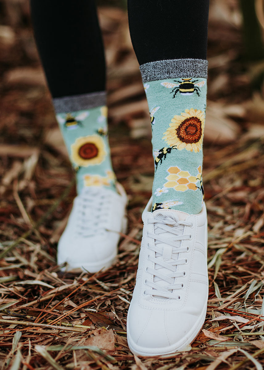 A female model wearing sunflower and bee-themed novelty socks and white sneakers poses in dry grass.