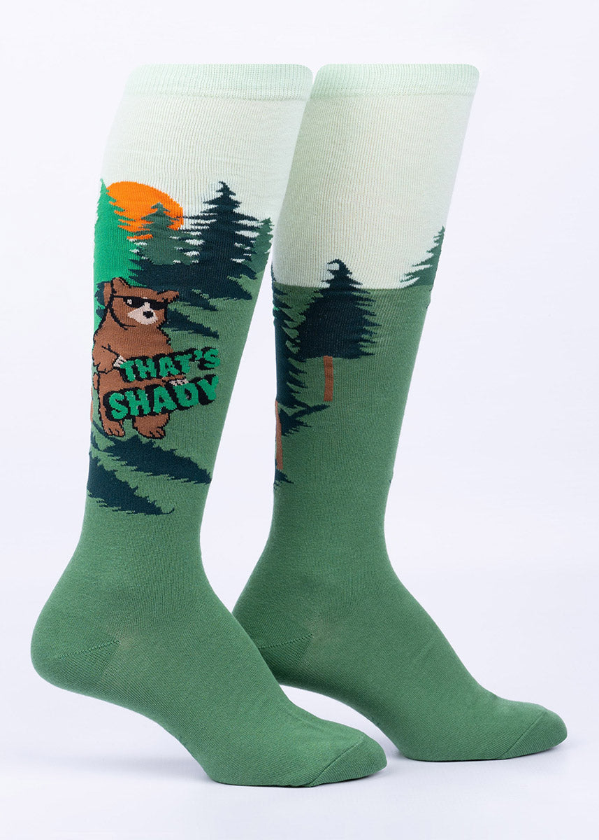 Green knee socks with a design featuring big brown grizzly bears walking upright while sporting black sunglasses as they turn their backs on the setting sun and hold up the words “That's Shady.” 