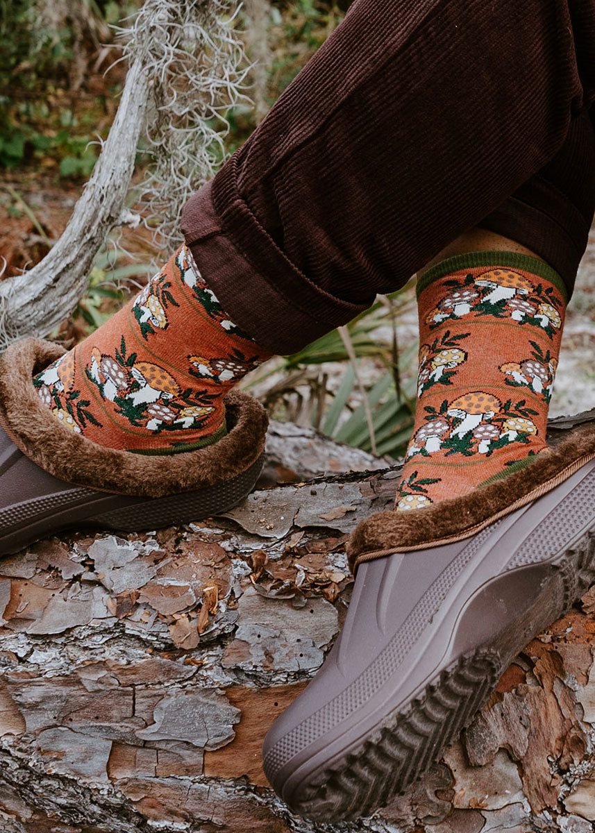 A model wearing mushroom-themed novelty socks poses outside on a wooden log.