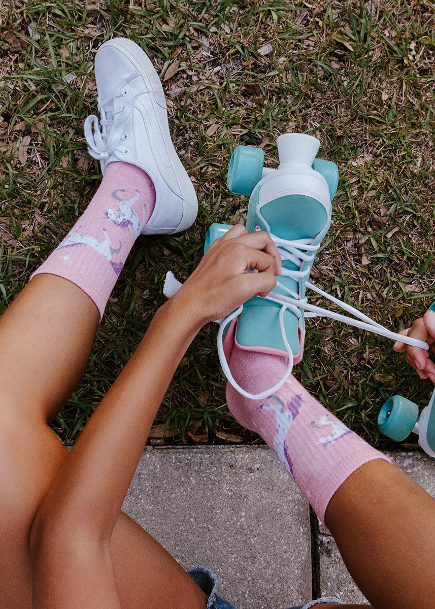 A model wearing pink unicorn-themed novelty socks poses while lacing up a blue rollerskate.
