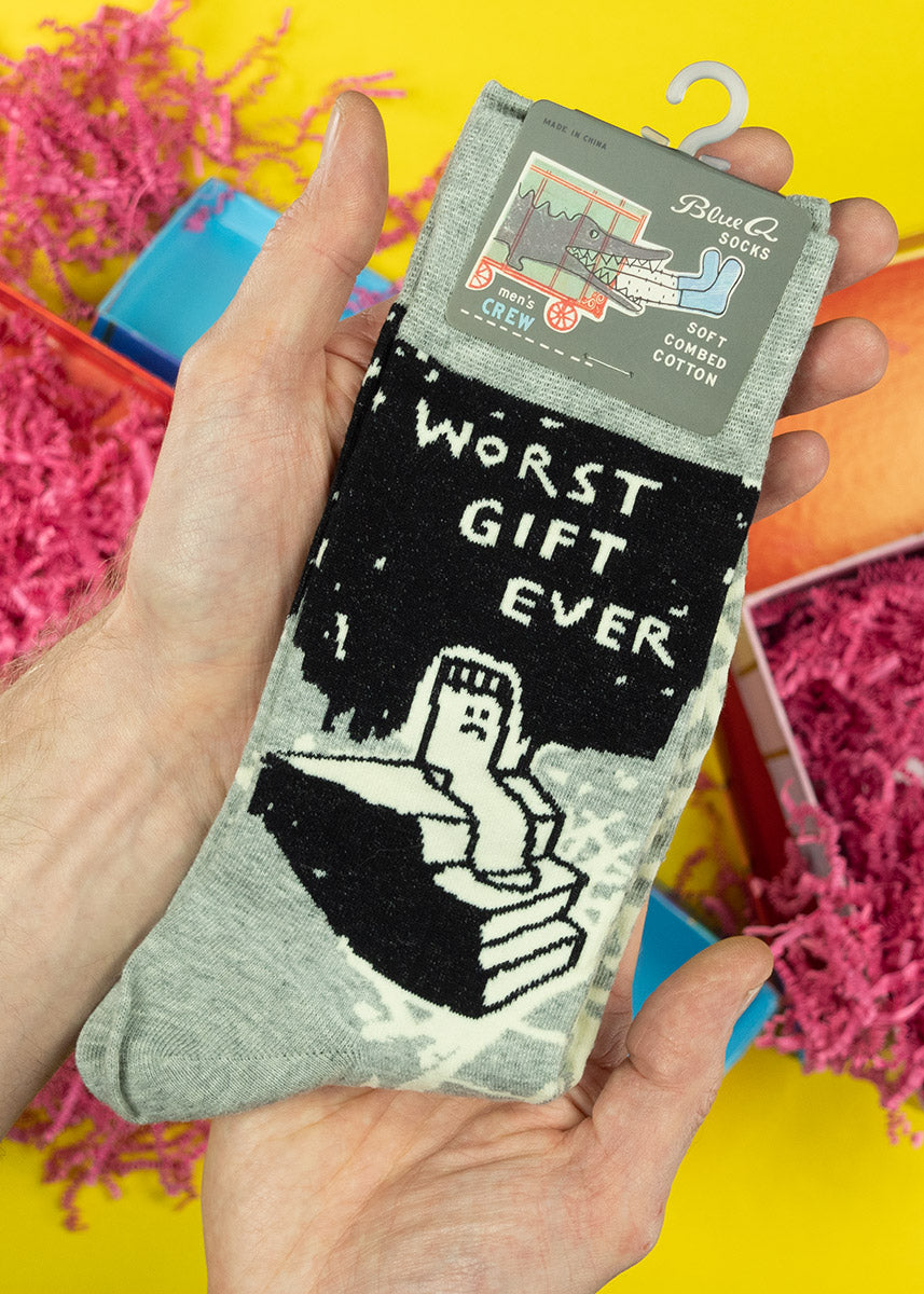 A model holds a pair of black, white, and grey socks that say &quot;Worst Gift Ever&quot; with a sad sock sitting on a set of stairs. 